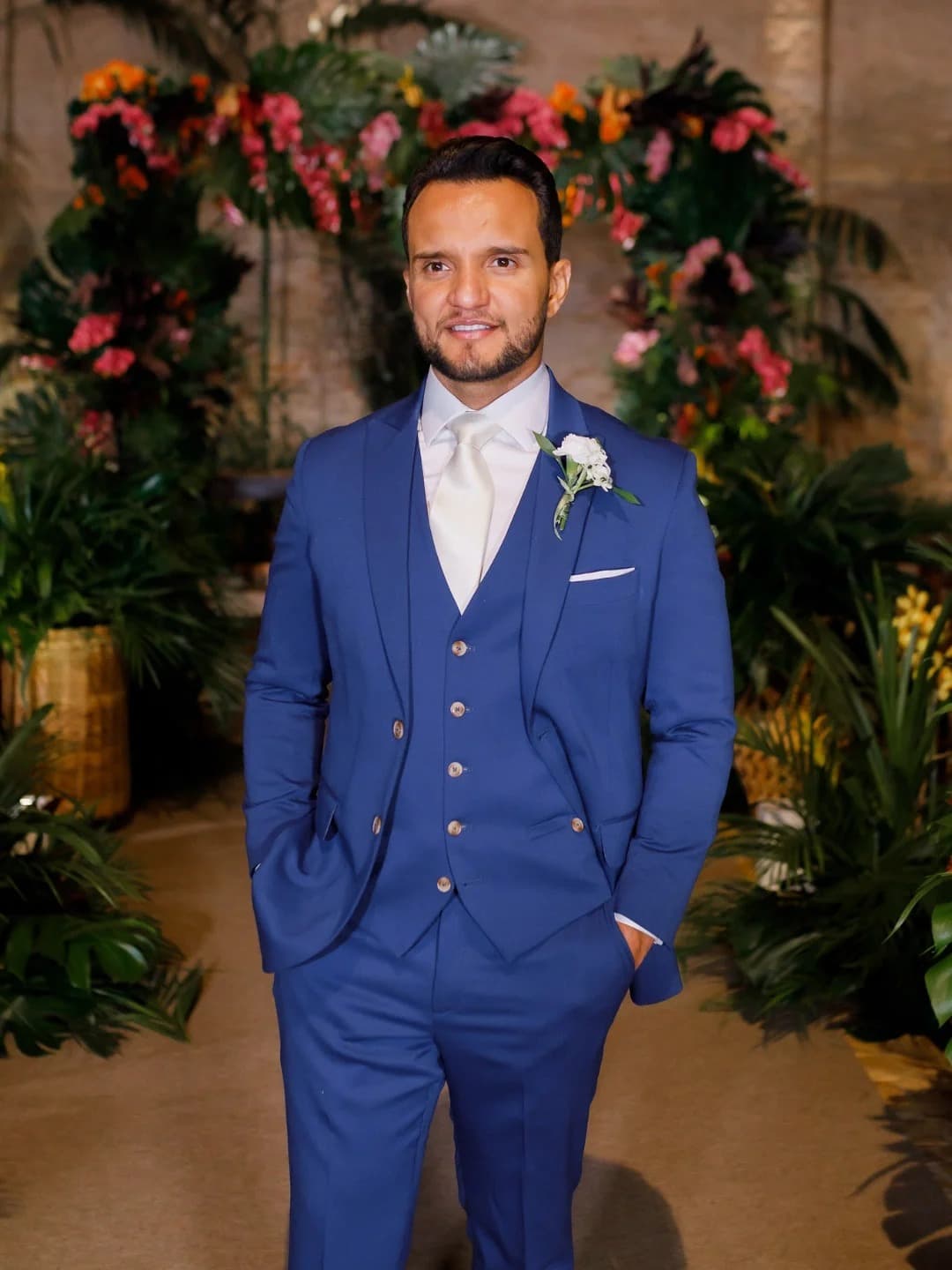 Noivo em terno azul-royal três peças diante de arco de flores